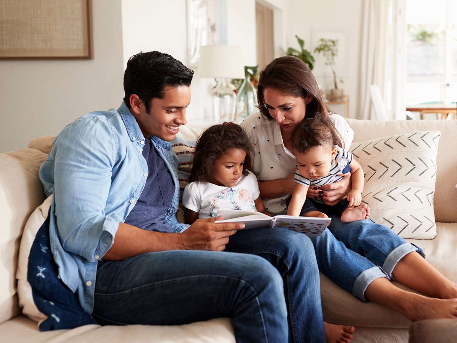 Hispanic couple sitting on the sofa reading a book at home with their baby son and young daughter