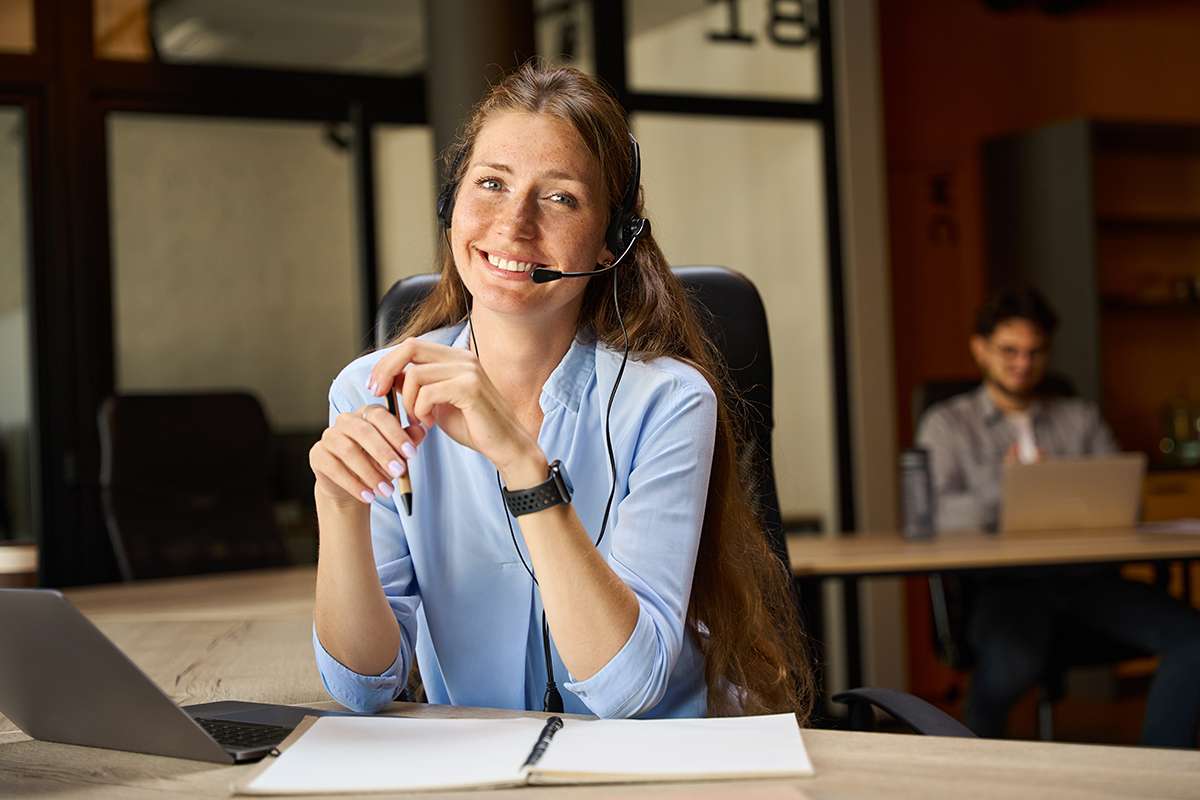 Smiling female call center operator looking at camera near blurred colleague.